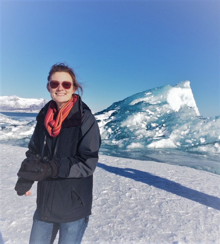 K Chess, a happy woman in sunglasses stands in front of an iceberg.